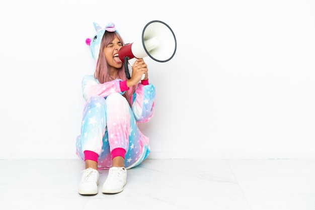 Young mixed race woman wearing a unicorn pajama sitting on the floor isolated on white background shouting through a megaphone