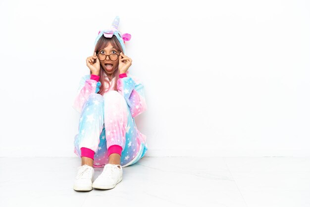 Young mixed race woman wearing a unicorn pajama sitting on the floor isolated on white background with glasses and surprised