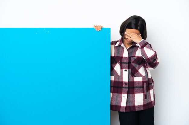 Young mixed race woman with a big blue placard isolated on white background making stop gesture and covering face