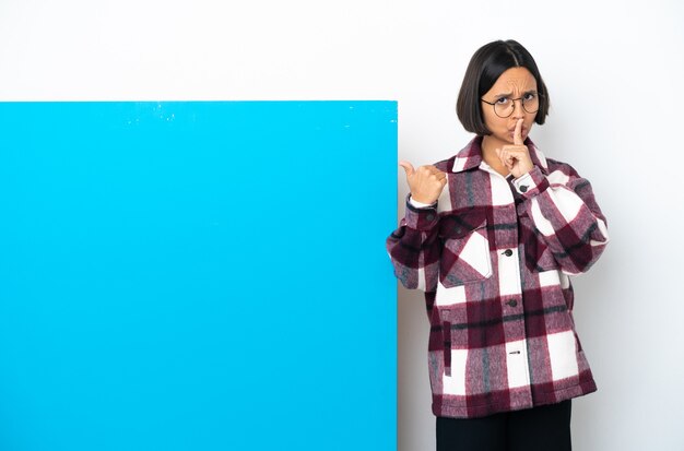 Молодая женщина смешанной расы с большим синим плакатом, изолированным на белом фоне, указывая в сторону и делая жест молчания