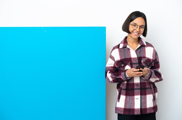 携帯電話でメッセージを送信する白い背景で隔離の大きな青いプラカードを持つ若い混血の女性