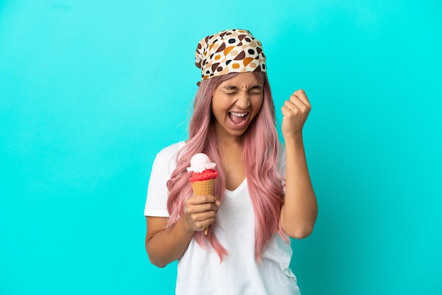 Giovane donna di razza mista con un gelato alla cornetta isolato su sfondo blu che celebra una vittoria