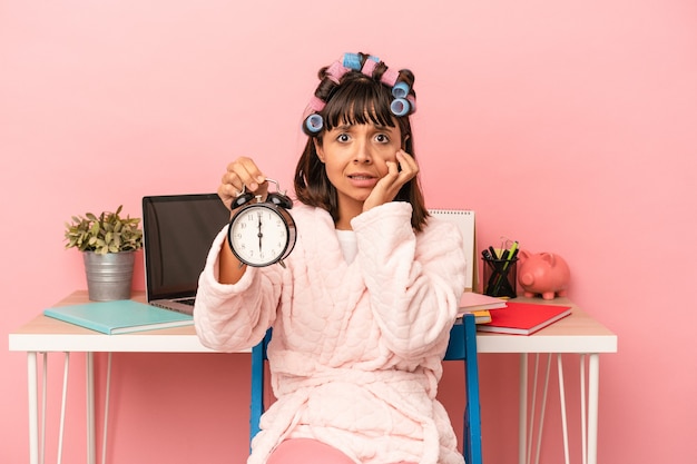 Photo young mixed race woman with curlers holding alarm clock isolated on pink background