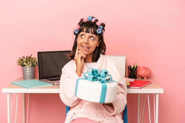 Young mixed race woman with curlers holding cake isolated on pink background