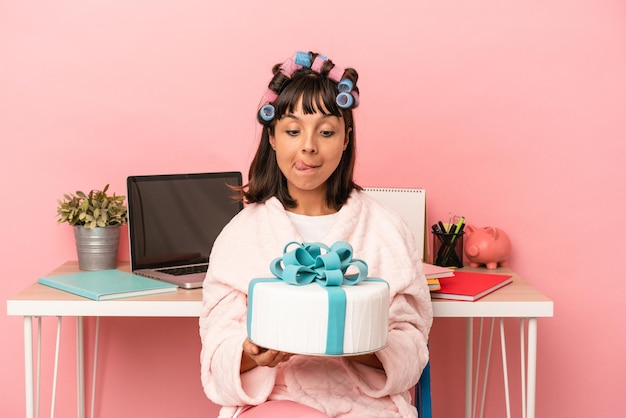 Young mixed race woman with curlers holding cake isolated on pink background