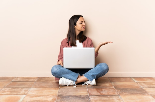 Young mixed race woman with a laptop sitting on the floor holding copyspace with doubts