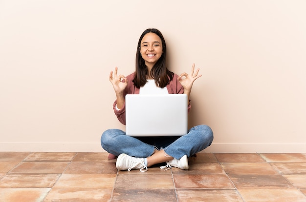 Young mixed race woman with a laptop sitting on the floor showing ok sign with two hands