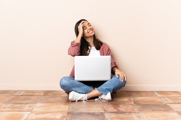 Young mixed race woman with a laptop sitting on the floor smiling a lot