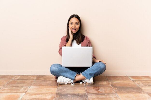 Young mixed race woman with a laptop sitting on the floor with surprise and shocked facial expression