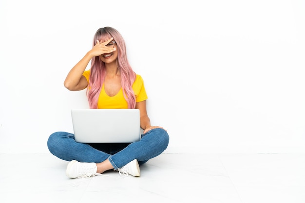 Photo young mixed race woman with a laptop with pink hair sitting on the floor isolated on white background covering eyes by hands and smiling