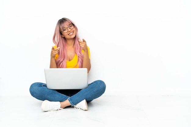 Young mixed race woman with a laptop with pink hair sitting on the floor isolated on white background points finger at you while smiling