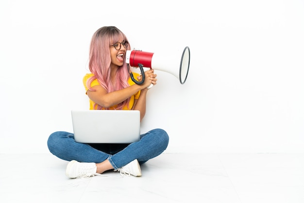 Молодая женщина смешанной расы с ноутбуком с розовыми волосами сидит на полу, изолированном на белом фоне, кричит в мегафон