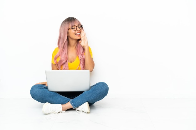 Молодая женщина смешанной расы с ноутбуком с розовыми волосами сидит на полу на белом фоне и кричит с широко открытым ртом в стороны