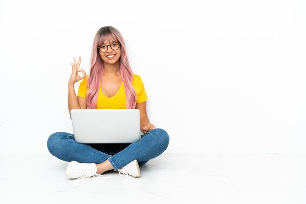 Young mixed race woman with a laptop with pink hair sitting on the floor isolated on white background showing ok sign with fingers