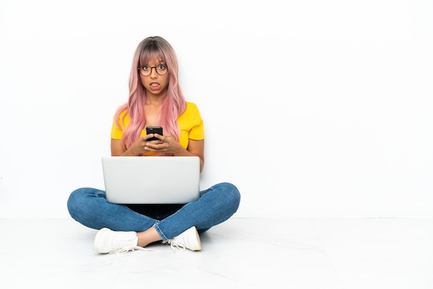 Foto giovane donna di razza mista con un computer portatile con i capelli rosa seduta sul pavimento isolato su sfondo bianco sorpresa e l'invio di un messaggio