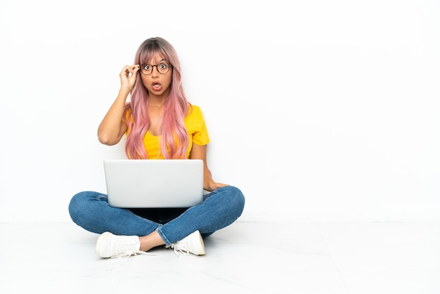 Young mixed race woman with a laptop with pink hair sitting on the floor isolated on white background with glasses and surprised