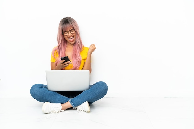 Foto giovane donna di razza mista con un computer portatile con i capelli rosa seduta sul pavimento isolato su sfondo bianco con il telefono in posizione di vittoria