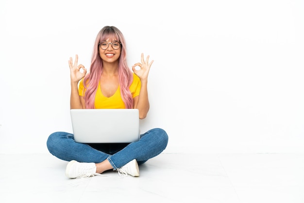 Young mixed race woman with a laptop with pink hair sitting on the floor isolated on white background in zen pose