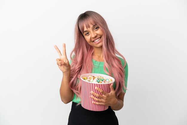 Young mixed race woman with pink hair eating popcorn isolated on white background smiling and showing victory sign