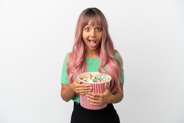 Young mixed race woman with pink hair eating popcorn isolated on white background with surprise facial expression