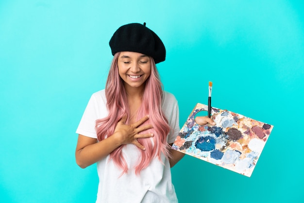 Фото Молодая женщина смешанной расы с розовыми волосами держит палитру, изолированную на синем фоне, много улыбаясь
