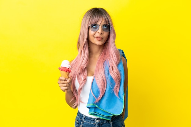 Young mixed race woman with pink hair holding ice cream isolated on yellow background and looking up