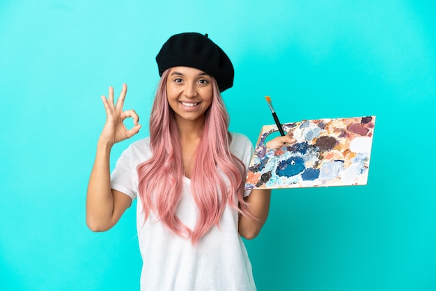 Foto giovane donna di razza mista con capelli rosa in possesso di una tavolozza isolata su sfondo blu che mostra segno ok con le dita