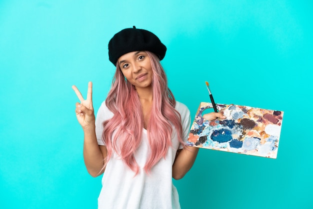 Young mixed race woman with pink hair holding a palette isolated on blue background smiling and showing victory sign