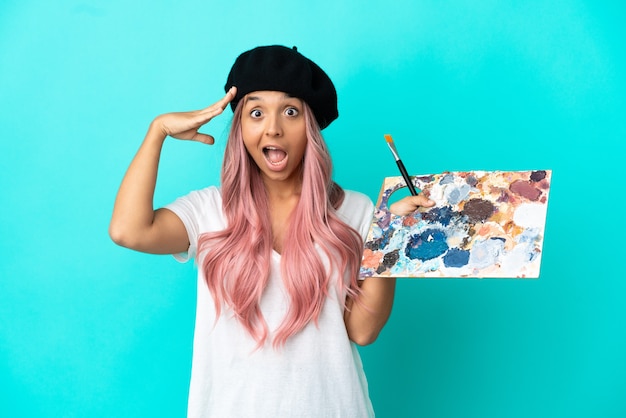 Young mixed race woman with pink hair holding a palette isolated on blue background with surprise expression