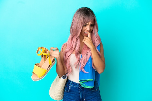 Молодая женщина смешанной расы с розовыми волосами держит летние сандалии, изолированные на синем фоне, сомневаясь