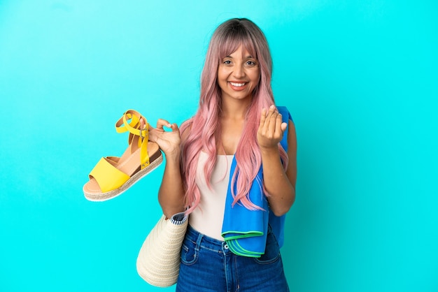 Young mixed race woman with pink hair holding summer sandals isolated on blue background inviting to come with hand. Happy that you came