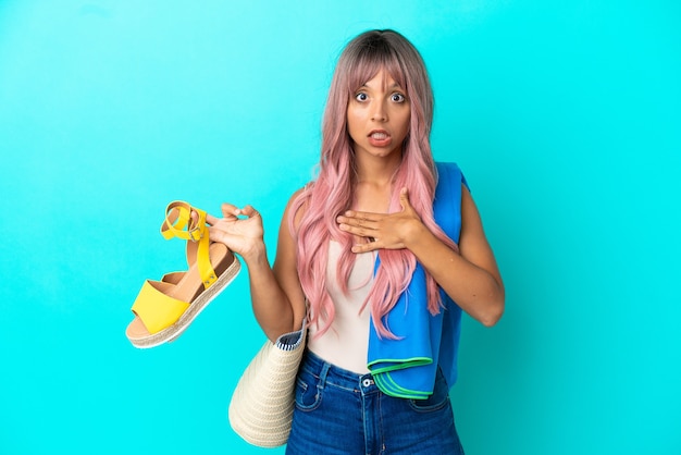 Young mixed race woman with pink hair holding summer sandals isolated on blue background surprised and shocked while looking right