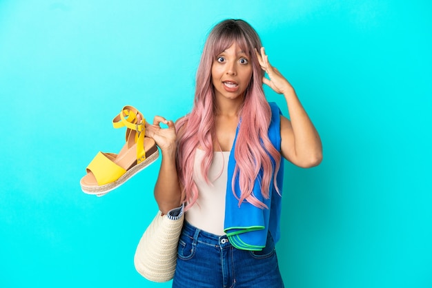 Young mixed race woman with pink hair holding summer sandals isolated on blue background with surprise expression