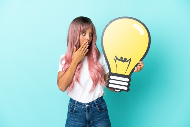 Молодая женщина смешанной расы с розовыми волосами изолирована на синем фоне, держа значок лампочки с удивленным выражением лица