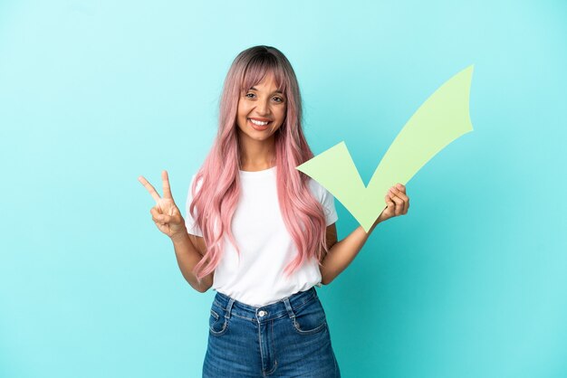 Young mixed race woman with pink hair isolated on blue background holding a check icon and celebrating a victory