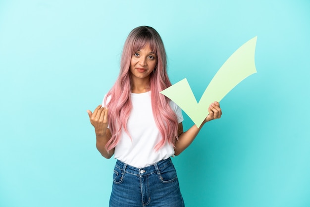 Young mixed race woman with pink hair isolated on blue background holding a check icon and doing coming gesture