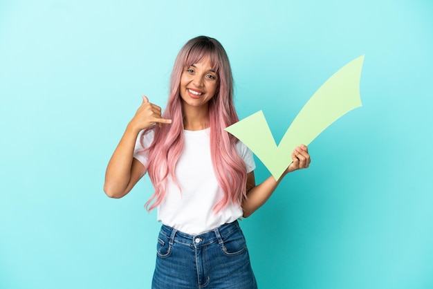 Young mixed race woman with pink hair isolated on blue background holding a check icon and doing phone gesture