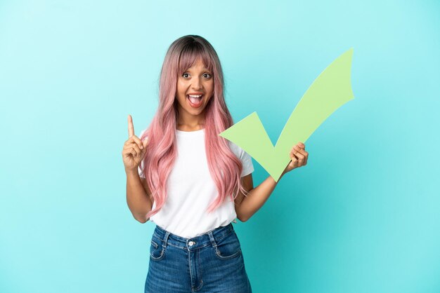 Young mixed race woman with pink hair isolated on blue background holding a check icon and pointing up