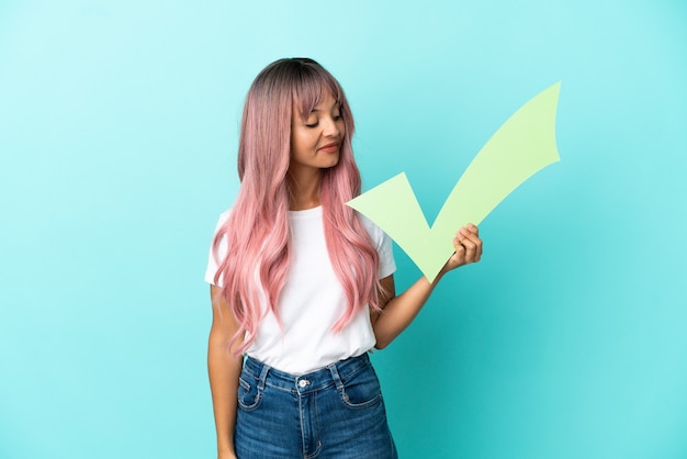 Young mixed race woman with pink hair isolated on blue background holding a check icon with happy expression