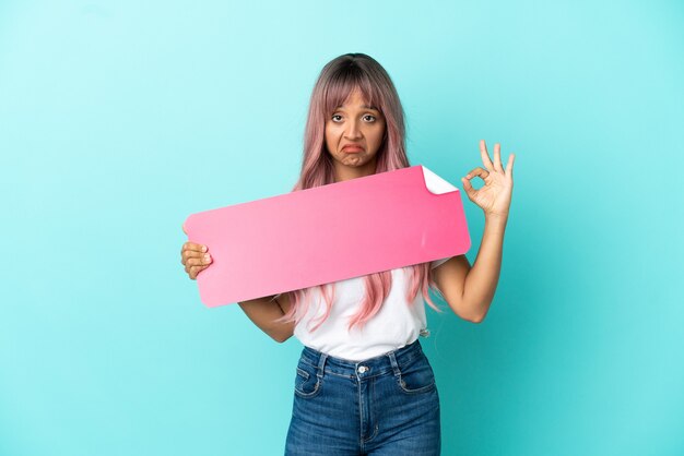 Young mixed race woman with pink hair isolated on blue background holding an empty placard and doing OK sign