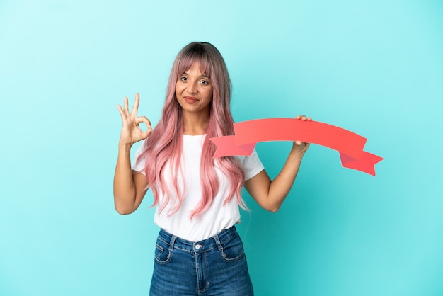 Giovane donna di razza mista con capelli rosa isolata su sfondo blu con in mano un cartello vuoto e facendo segno ok