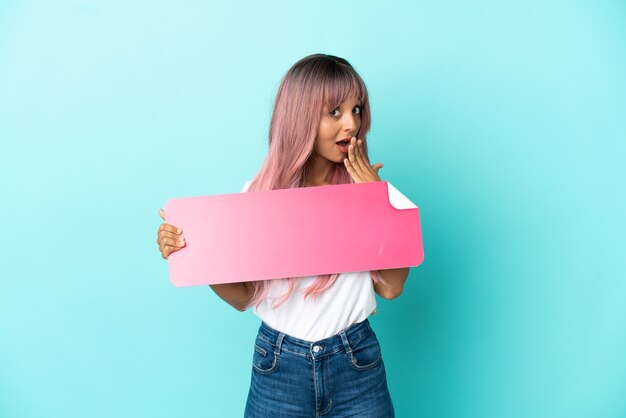 Young mixed race woman with pink hair isolated on blue background holding an empty placard with surprised expression