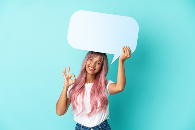 Young mixed race woman with pink hair isolated on blue background holding an empty speech bubble and doing OK sign