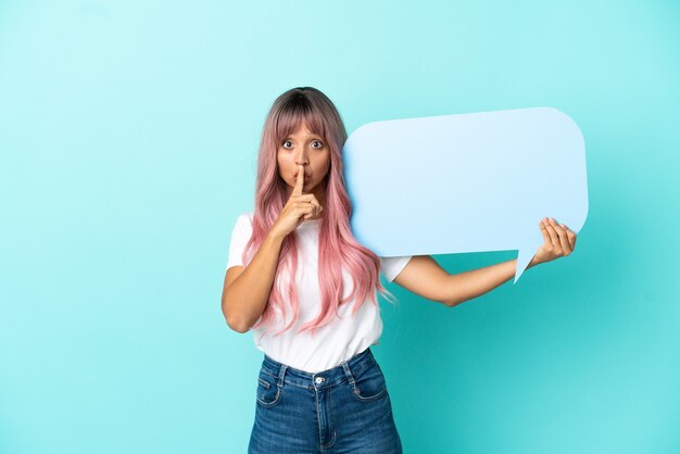 Young mixed race woman with pink hair isolated on blue background holding an empty speech bubble and doing silence gesture