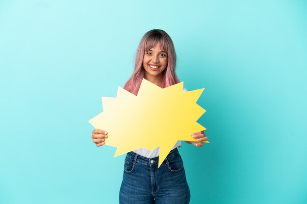 Young mixed race woman with pink hair isolated on blue background holding an empty speech bubble