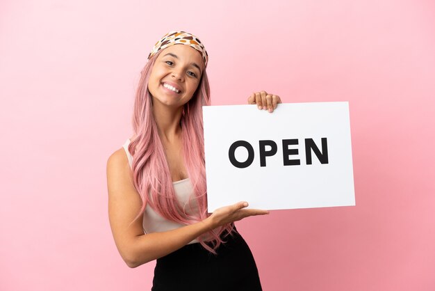 Young mixed race woman with pink hair isolated on pink background holding a placard with peace symbol with happy expression