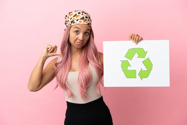 Young mixed race woman with pink hair isolated on pink background holding a placard with recycle icon with proud gesture