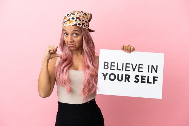ピンクの背景に分離されたピンクの髪を持つ若い混血の女性は、誇らしげなジェスチャーであなたの自己を信じてテキストのプラカードを保持しています