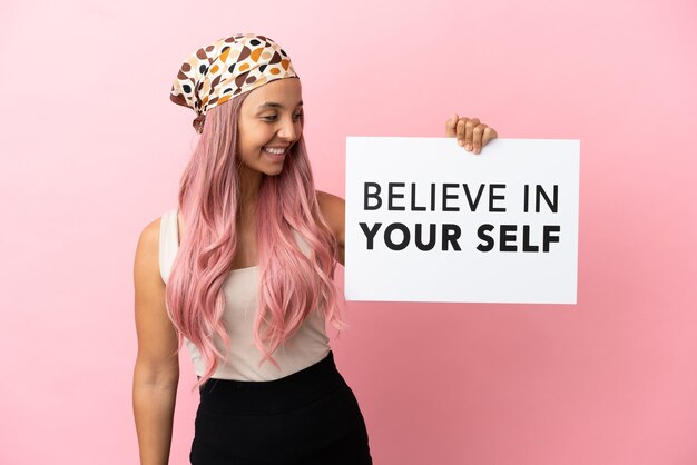 Photo young mixed race woman with pink hair isolated on pink background holding a placard with text believe in your self