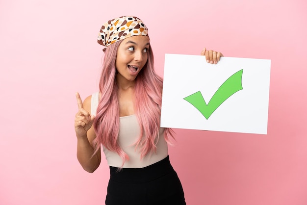 Young mixed race woman with pink hair isolated on pink background holding a placard with text Green check mark icon and thinking